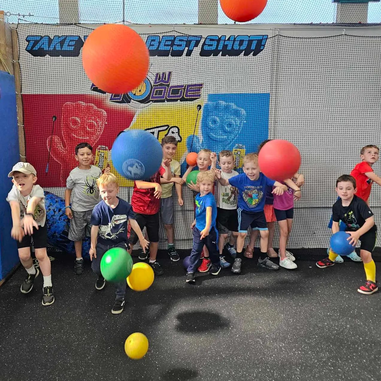 A group of children stand and play with colorful balls in an indoor play area, smiling and mid-action, with a dodgeball-themed backdrop behind them that reads "TAKE YOUR BEST SHOT!.