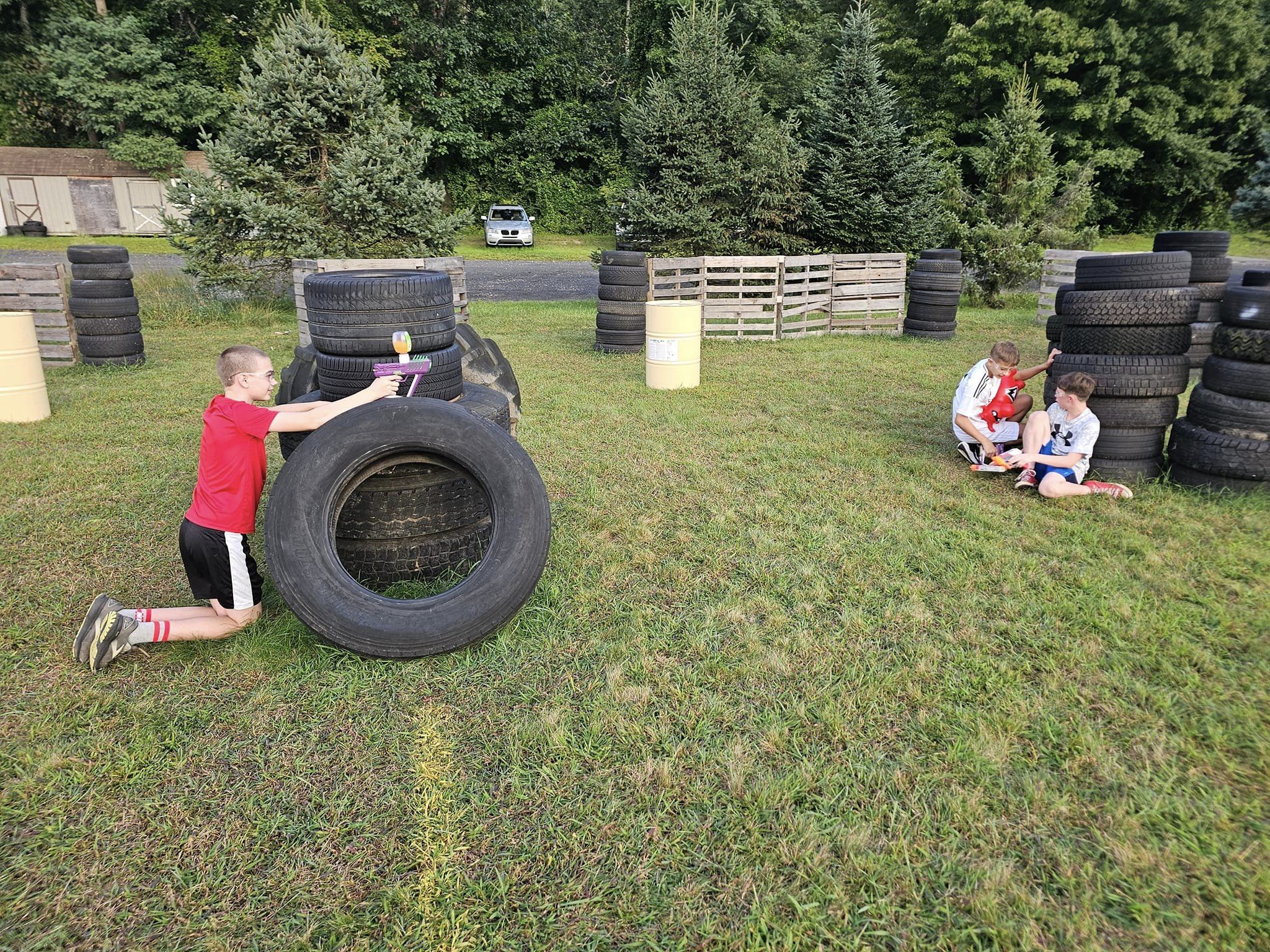 A boy in a red shirt hides behind large tires aiming a toy blaster, while three boys in white and red shirts sit together near more tires on a grassy field, with trees and barrels in the background.