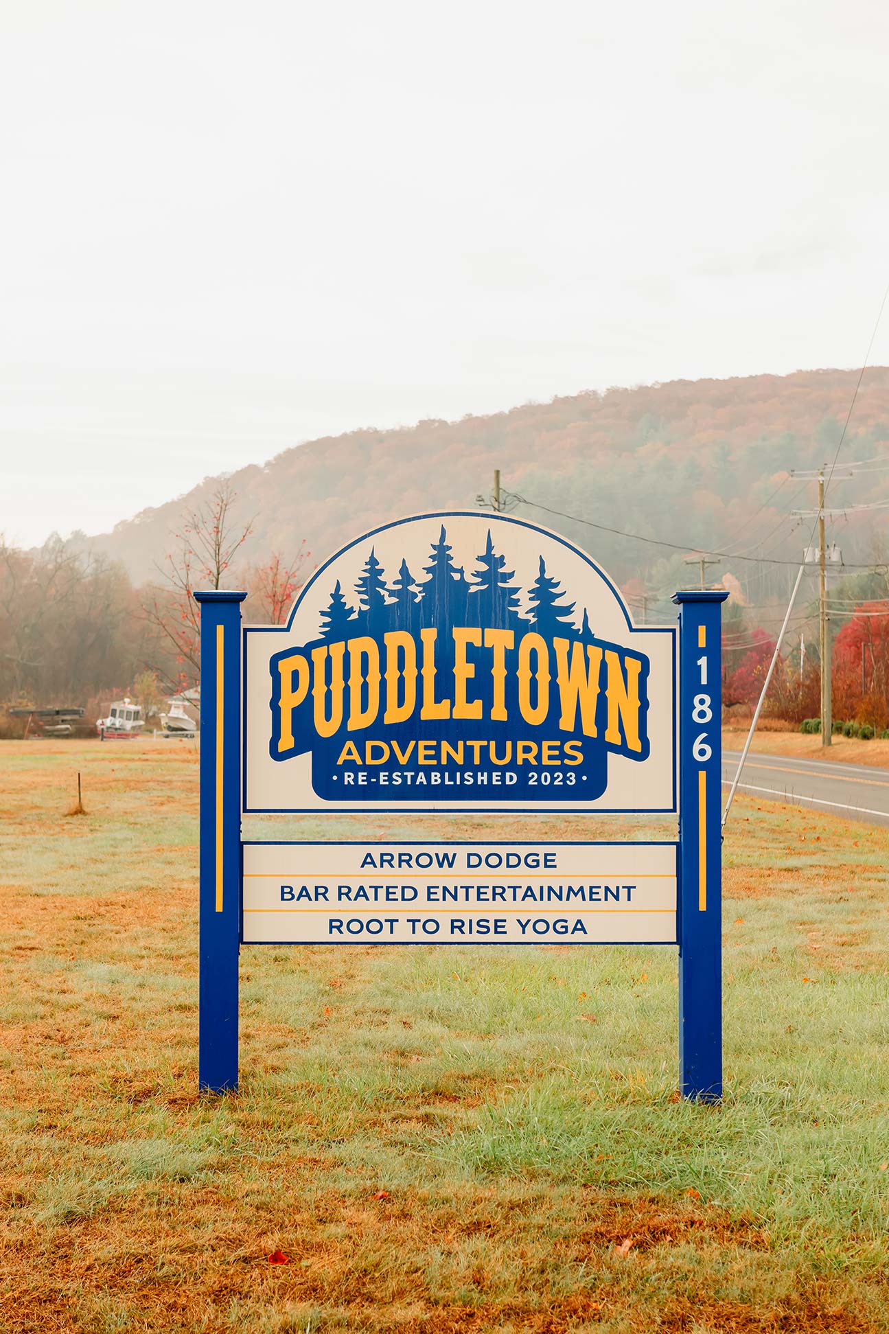 A sign for Puddletown Adventures reads: “Puddletown Adventures Re-Established 2023. Arrow Dodge. Bar Rated Entertainment. Root to Rise Yoga.” The sign stands on grass near a road with hills and trees in the background.