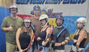 Six adults wearing helmets and holding bows pose together in front of a netted backdrop with colorful signage. They appear to be participating in an indoor archery or combat archery activity.