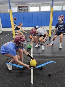 Four teens in helmets and masks, each holding a bow, crouch and compete for rubber balls in a gym. They appear to be playing a team activity involving archery and soft balls. Blue mats and yellow poles are visible in the background.