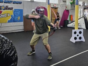 A man wearing a helmet and face shield aims a bow and arrow indoors. Other people and archery targets are in the background, and a banner on the wall reads "Dodge.