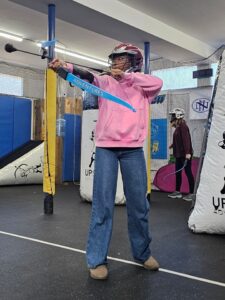 A person wearing a helmet and face shield aims a blue bow indoors. They are dressed in a pink hoodie and jeans. Inflatable barriers and another helmeted person are visible in the background.