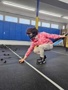 A person in a pink camouflage hoodie, gray pants, and sneakers crouches in a gym, wearing a blindfold and reaching down to touch several cords or ropes on the floor. Blue wall padding and yellow posts are in the background.