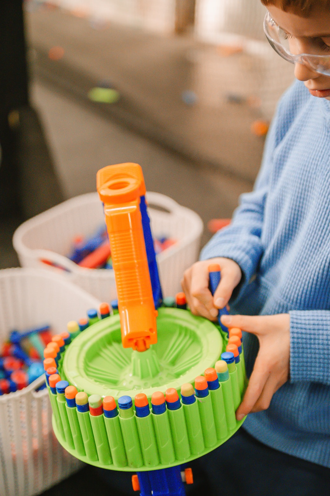 A child wearing safety glasses and a blue sweater loads foam darts into a large green toy blaster drum magazine, with bins of more toy darts nearby.
