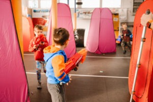 Three young boys play with large toy Nerf blasters in an indoor arena with colorful inflatable barriers. The scene is active and playful, suggesting a friendly Nerf gun battle.