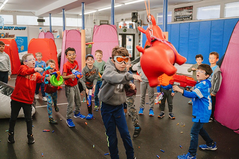 A group of children excitedly watch as one boy wearing goggles tries to hit a red inflatable animal piñata indoors. The kids are dressed casually and seem to be celebrating at a party.