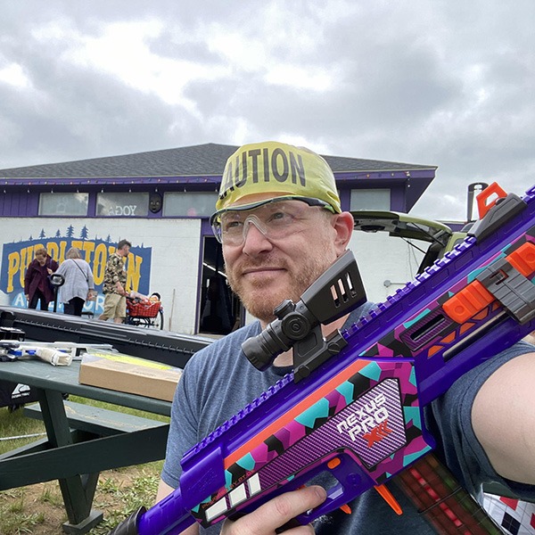 A man wearing safety glasses and a yellow "Caution" headband holds a colorful toy blaster outside near a picnic table, with a Purple Toad sign and other people in the background under a cloudy sky.