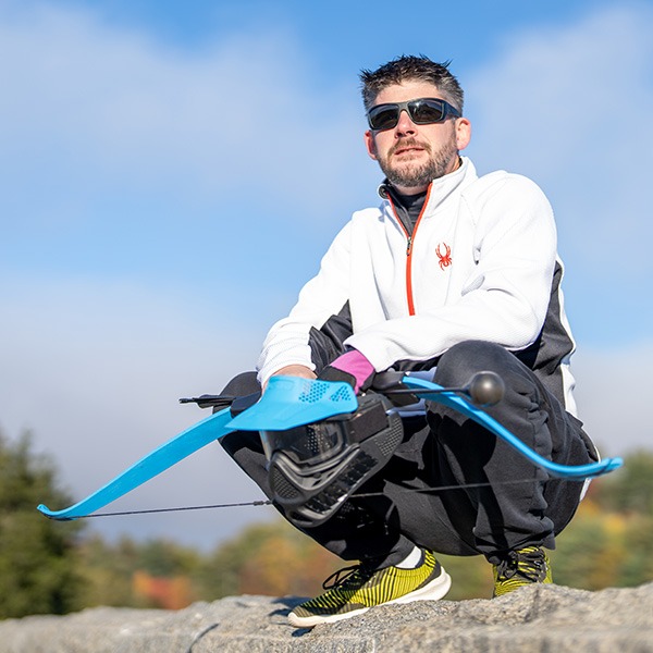 A man wearing sunglasses, a white jacket, and yellow running shoes crouches outdoors on a rocky surface, holding a blue and black adaptive bow in sunny weather. Trees and a blue sky are in the background.