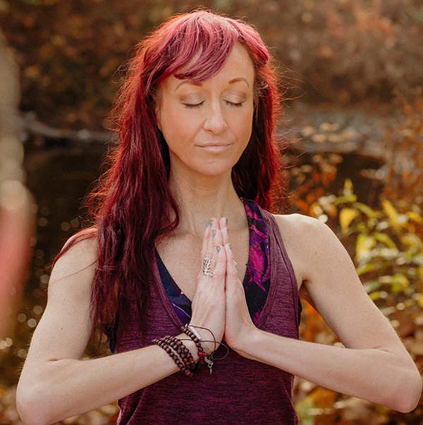 A woman with long red hair and closed eyes stands outdoors, hands pressed together in a prayer position, wearing a purple tank top and beaded bracelets, surrounded by blurred autumn foliage.
